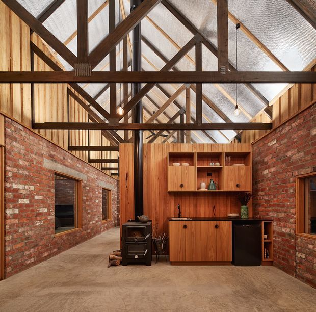 A rustic industrial interior with exposed red brick walls, timber wall panelling and cabinetry, and a dramatic high pitched roof structure. Dark timber trusses and rafters are fully exposed beneath corrugated translucent roof sheeting, creating a barn-like volume. The compact kitchen features warm timber joinery, open shelving, a dark benchtop, and a small black refrigerator. Concrete flooring and simple pendant lights reinforce the utilitarian character.