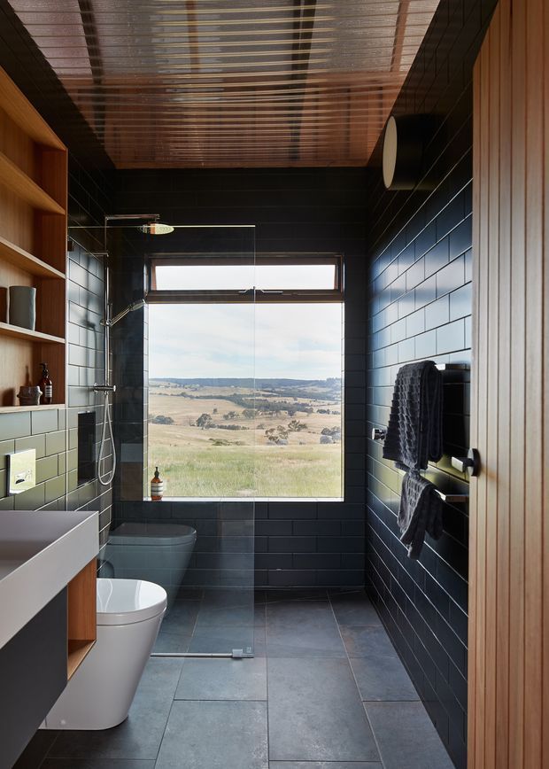 A narrow, modern bathroom viewed from the entry, featuring dark charcoal rectangular wall tiles, large grey floor tiles, a white freestanding bathtub and toilet, timber vanity shelving, and a glass-enclosed shower. A large rear window provides expansive rural landscape views. The ceiling has a distinctive linear timber or slatted treatment, while timber wall panelling frames the right side.