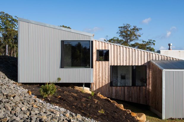 Exterior view of a contemporary, angular single-storey building on a sloped, rocky site. The facade combines vertically ribbed metal cladding with warm vertical timber boards, large dark-framed windows, and a pitched or folded roof form. Native vegetation and exposed rock surround the structure under a clear blue sky.