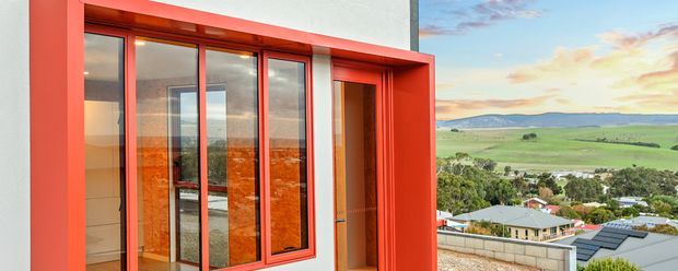 Exterior view of a contemporary multi-panel glazed facade and recessed entry framed in vivid red, with reflections of the surrounding landscape in the glazing. The building overlooks a suburban settlement and expansive green rolling countryside.