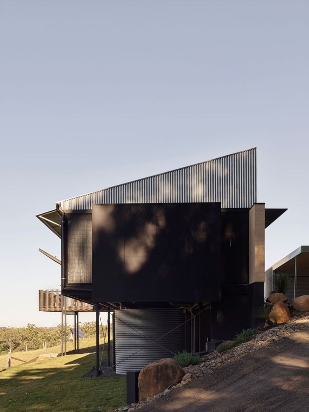 Exterior view of a contemporary, elevated house on a sloping site. The building has a dark angular metal-clad upper volume, corrugated metal roofing or screening, slender supports, projecting roof planes, a lower corrugated garage-like volume, timber balcony screening, and exposed rock landscaping.