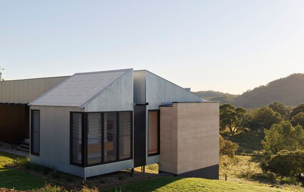 Exterior view of a contemporary single-storey house on a sloping, vegetated site. The building features corrugated metal cladding and roofing, a projecting gabled volume with large dark-framed glazed openings, and a smooth pale rendered or concrete wall element. Tree-covered hills are visible in the background.