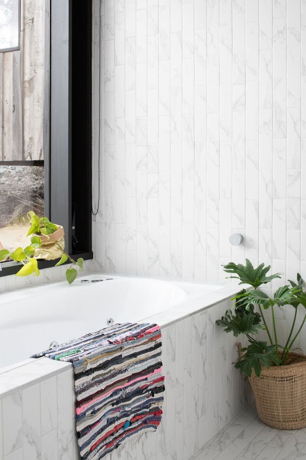 Bright bathroom featuring a white built-in bathtub enclosed by white marble-look rectangular wall tiles and matching tiled bath surround. A black-framed window provides natural light, with potted green plants and a colourful woven textile adding contrast. The room has a fresh, clean, relaxed character.