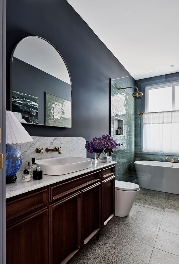 A sophisticated bathroom with a dark charcoal painted feature wall, arched mirror, marble vanity top, dark timber cabinetry, white vessel sink, and brass fixtures. Green glazed subway tiles line the shower wall, with a freestanding bathtub beneath a large window. Grey tiled flooring and refined decorative details create a moody, elegant interior.