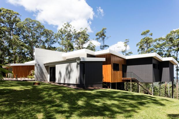 Exterior view of a contemporary single-storey house set among established trees on a gently sloping grassy site. The home features intersecting flat roof forms, white rendered walls, dark vertical cladding, warm timber cladding, broad glazing, and a raised deck or entry stair.