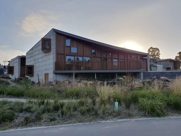 Exterior view of a contemporary multi-level residential building with a long dark timber-clad upper facade, exposed concrete side walls, broad balcony, and a low-pitched asymmetrical roof. The building sits within a landscaped streetscape with native grasses and a paved road in the foreground.