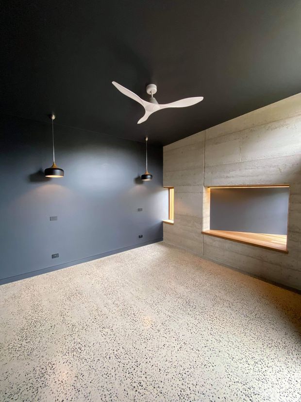 An empty, dark-toned contemporary interior with a polished terrazzo floor, charcoal painted walls and ceiling, and a large exposed concrete feature wall. The concrete wall includes a recessed timber-lined display or serving niche with concealed warm lighting, while two black pendant lights and a sculptural white ceiling fan add contrast. A narrow timber-framed opening provides additional natural light.