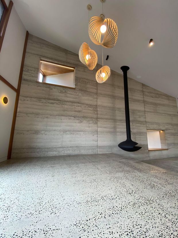 A minimalist interior with a large polished terrazzo floor and exposed horizontal-textured concrete wall panels. Three sculptural woven pendant lights hang from the white ceiling, while a black suspended wood-burning fireplace provides a strong vertical feature. Timber-framed recessed openings and warm timber detailing add contrast to the neutral concrete palette.