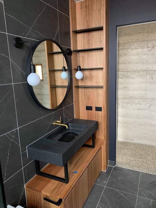 Contemporary bathroom vanity area featuring a black stone benchtop with integrated basin, brass wall-mounted tapware, round black-framed mirror, globe sconces, dark veined wall tiles, and warm timber shelving and cabinetry. A polished concrete-like surface is visible through the doorway.