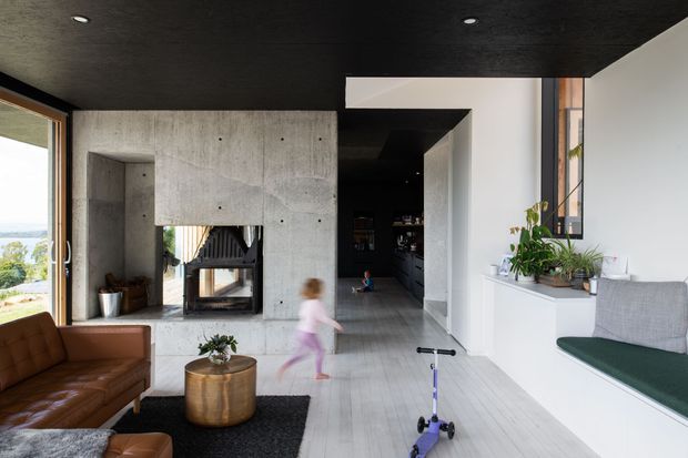 Open-plan contemporary living area with a dark textured ceiling, exposed concrete fireplace surround, pale timber flooring, and a built-in window seat. A brown leather sofa and round metallic coffee table sit beside the fireplace, while a dark kitchen or bar area recedes into the background. Large glazing provides a view toward the landscape, and the room includes indoor plants and children's toys.
