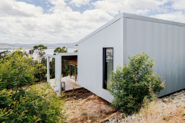 Exterior view of a compact contemporary building set on a sloped, vegetated site. The structure has crisp geometric forms, pale grey vertically ribbed metal cladding, a low-pitched roofline, dark-framed narrow glazing, and a covered timber deck or entry recess. Native-looking vegetation surrounds the building, with distant hills visible beyond.