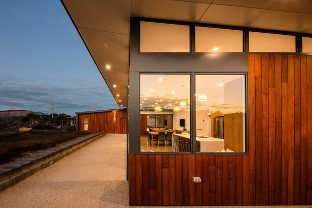 Exterior view of a contemporary single-storey building at dusk, featuring warm vertical timber wall cladding, dark-framed glazing, clerestory windows, and a broad projecting roof with recessed lighting. A paved walkway leads past the facade, while the windows reveal a brightly lit dining and kitchen area inside.