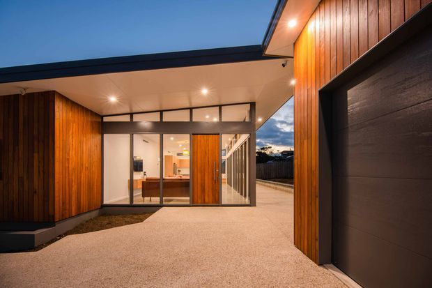 Twilight exterior view of a contemporary residential entry and garage. The home features warm vertical timber wall cladding, dark-framed glazed entry doors and clerestory glazing, a broad flat roof overhang with recessed downlights, and a dark sectional garage door. A light aggregate driveway leads through the covered entry toward a warmly lit interior.
