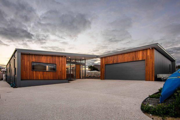 A contemporary single-storey building exterior viewed from a broad paved driveway. The composition features two dark-clad wings with warm vertical timber-look wall cladding, low-pitched gabled roof forms, a glazed central entry connection, and a large dark sectional garage door. The setting includes minimal landscaping and an overcast sky.