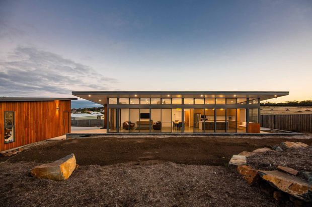 A low, single-storey contemporary home viewed from the landscaped rear yard at dusk. The main pavilion has a broad flat roof with deep overhangs and an almost fully glazed facade, revealing a warmly lit open-plan interior. A separate timber-clad structure sits to the left, while exposed earth, large rocks, and timber fencing form the foreground and site boundary.