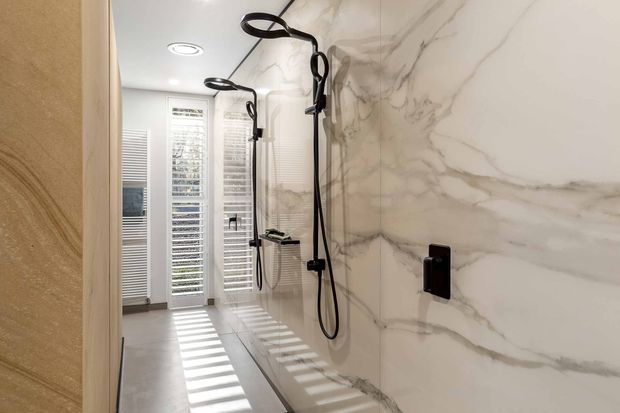 A bright, narrow contemporary bathroom with a walk-in double shower. Large white marble-look wall panels with grey veining dominate the right wall, paired with pale tiled flooring and matte black shower hardware. Plantation shutters at the far end cast striped sunlight across the floor, while recessed ceiling lighting provides additional illumination.