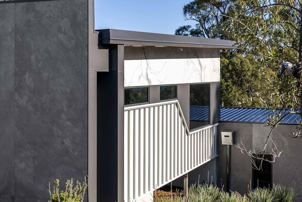 Exterior view of a contemporary multi-level building with a flat roof, dark textured wall cladding, contrasting white marble-look feature panels, dark-framed horizontal windows, and a white vertical-bar stair or balcony balustrade. A blue-grey metal roof is visible on the adjoining lower section, with established trees and planting around the property.