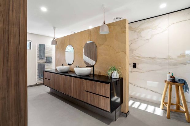 Contemporary bathroom featuring a long double vanity with dark timber cabinetry, black benchtop, two vessel basins and rounded mirrors. A warm timber partition separates the vanity from a walk-in shower lined in large white marble-look slabs. Neutral grey flooring, pendant lights and minimal black detailing create a refined, modern scheme.