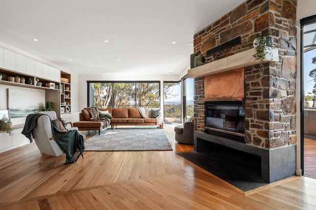 A spacious, warm living room with a substantial exposed stone fireplace and timber mantel as the focal point. The room features herringbone timber flooring, built-in white shelving and cabinetry, a neutral sectional sofa, lounge chairs, a large rug, and extensive windows overlooking trees and a distant landscape. The open-plan space is bright and inviting, with a contemporary fit-out balanced by rustic natural materials.
