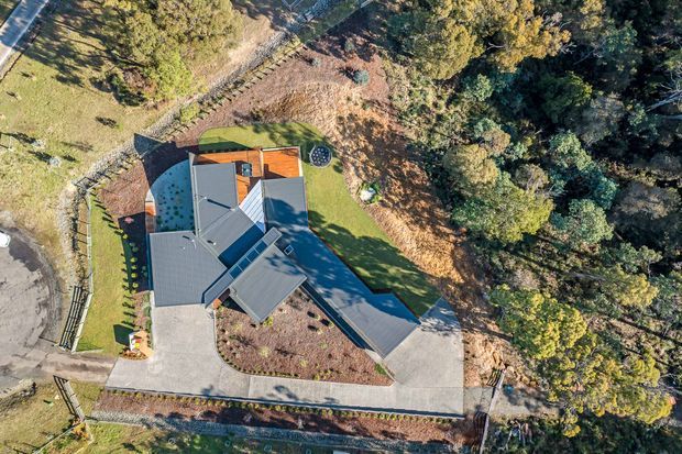 Aerial view of a detached residential property set within a mature, heavily landscaped site. The building has a complex multi-wing form with several pitched roof sections in grey metal, a small timber-toned deck or terrace, and broad pale paved driveways or paths surrounding the house. Established trees, lawn, garden beds, and boundary fencing dominate the surrounding grounds.