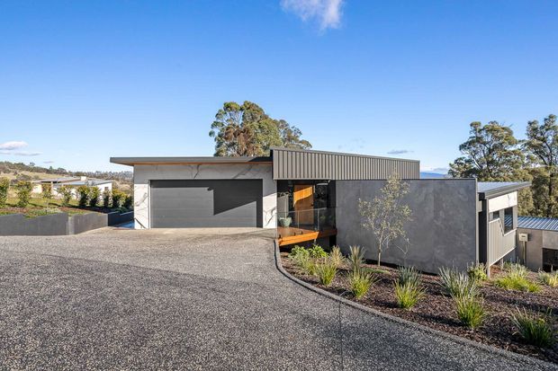 Exterior view of a contemporary single-storey residence on a sloping site, featuring a broad dark gravel driveway, charcoal rendered walls, vertical corrugated metal cladding, a low-pitched roof, and a large integrated garage. Native-style landscaped planting borders the building, with surrounding trees and distant hills visible beyond.