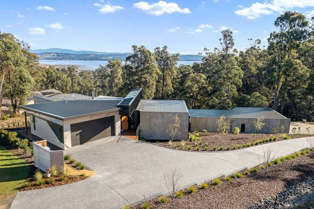 Aerial or elevated view of a contemporary low-rise residential property set among mature trees on a sloping site. The complex comprises several connected or closely grouped buildings with dark grey metal pitched roofs, grey rendered or concrete-like walls, a double garage, and a broad concrete driveway. A body of water and distant shoreline are visible beyond the tree canopy.