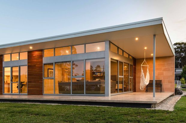 Single-storey contemporary house exterior at dusk, featuring a broad low-pitched roof with deep eaves, extensive floor-to-ceiling glazing in dark metal frames, warm timber wall cladding, and pale masonry feature walls. A covered terrace runs along the façade with a hanging chair and outdoor furniture, while warm interior lighting is visible through the windows.