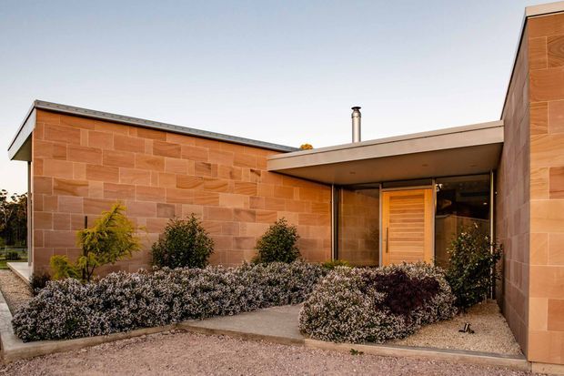 Single-storey contemporary building exterior with low-pitched roof forms, extensive warm terracotta-toned stone or masonry cladding, and a recessed entry beneath a broad flat canopy. Timber-toned entrance door and glazing sit within the sheltered entry court, bordered by drought-tolerant shrubs and gravel landscaping. The composition emphasizes strong horizontal lines, planar walls, and an understated modern aesthetic.