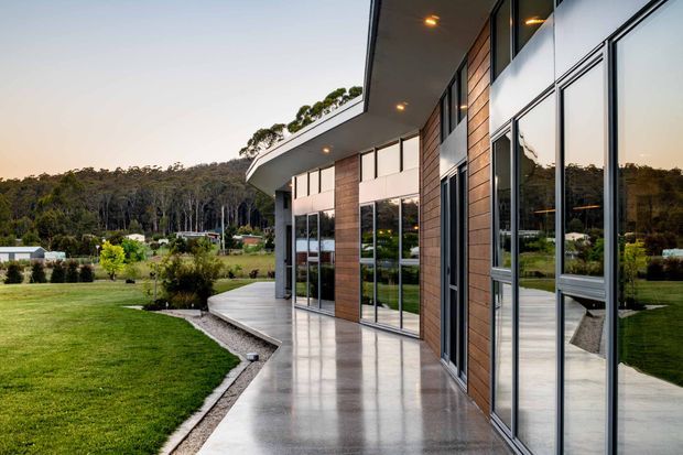 Contemporary single-storey building exterior viewed along a wide polished concrete verandah or terrace. The facade combines large floor-to-ceiling glazed panels with dark metal framing, warm vertical timber-clad sections, and a projecting flat roof with recessed downlights. The building overlooks a manicured lawn and landscaped rural garden, with wooded hills in the background.