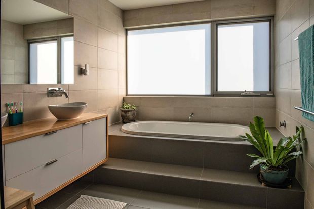 Contemporary bathroom with a built-in freestanding-style bathtub set into a tiled platform, a timber-topped floating vanity with vessel basin, large windows, and neutral stone-look wall and floor tiles. A mirror, wall-mounted fittings, towel rail, and potted plant complete the clean, warm-toned interior.