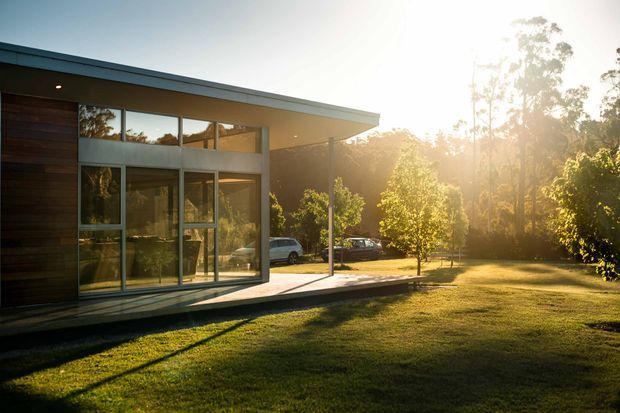 A low-profile modern house set within a leafy landscaped site, photographed at sunset. The building features a broad flat roof with deep eaves, extensive floor-to-ceiling glazing, dark horizontal timber cladding, slim light-coloured framing, and a covered terrace or deck extending toward the lawn. Warm sunlight and surrounding trees create a serene indoor-outdoor atmosphere.