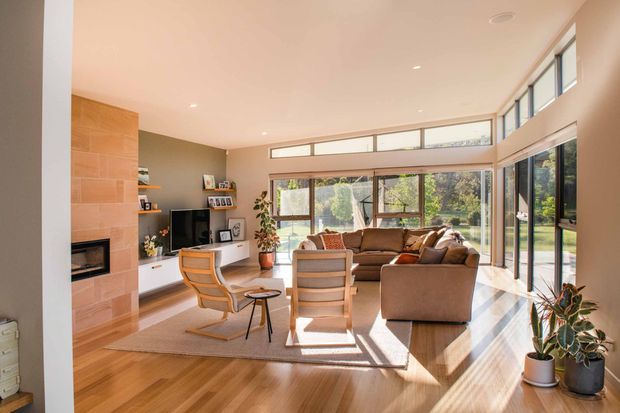 Bright contemporary living room with warm timber flooring, a large sectional sofa, lounge chairs, built-in shelving and television cabinetry, and a tiled fireplace feature wall. Floor-to-ceiling glazing and high clerestory windows bring in abundant natural light and overlook greenery. The open, airy room has a standard-to-high ceiling and a calm, refined neutral palette.