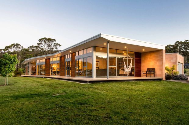 Exterior view of a contemporary single-storey house set within a spacious lawn and landscaped suburban or semi-rural site. The building has a long low-profile form, a prominent flat roof with deep covered outdoor areas, extensive floor-to-ceiling glazing, exposed warm timber-toned wall sections, and a large light brick masonry volume. Warm interior lighting is visible through the windows at dusk.