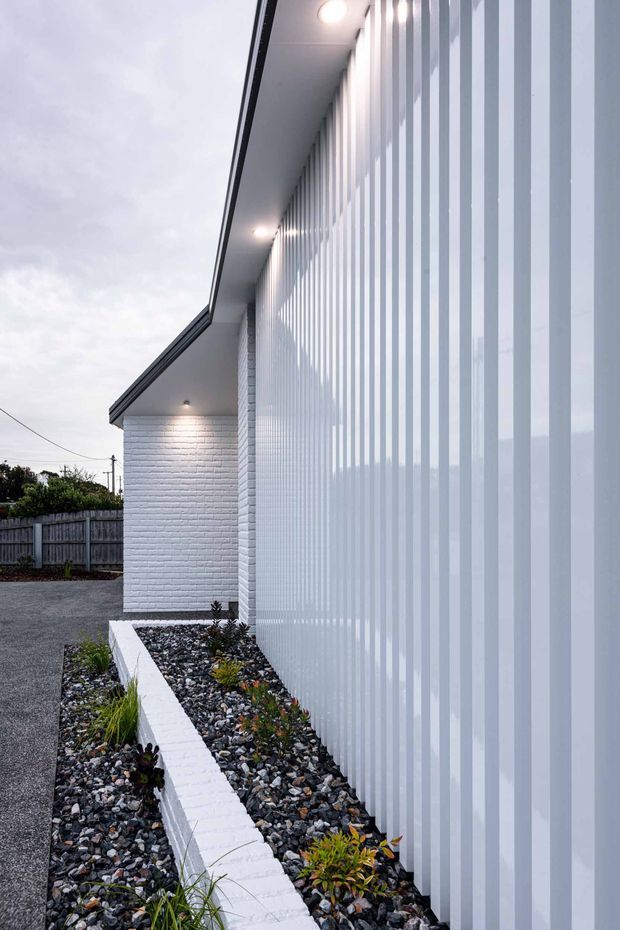 Photograph of a contemporary single-storey building exterior, featuring a long translucent vertically ribbed facade screen, white painted brick wall, deep roof eaves with recessed downlights, and a narrow landscaped gravel bed edged by a low white brick planter wall. An asphalt driveway and timber boundary fence are visible alongside the building.
