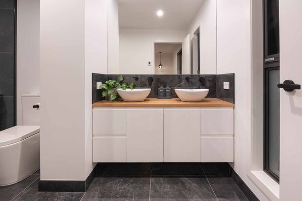 Contemporary bathroom vanity with a double-basin timber benchtop, white fixed cabinetry, dark stone-look tiled flooring and splashback, large mirror, and black-framed window. A toilet is visible in a partially separated alcove.