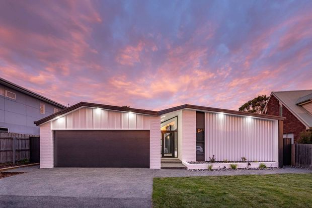 Contemporary single-storey suburban residence photographed from the street at dusk. The composition is dominated by a broad garage and white vertically ribbed facade, with a recessed glazed entry and a low-pitched roofline. A paved driveway, narrow landscaped garden strip, lawn, timber fencing, and neighbouring homes are visible. The dramatic pink and blue sunset sky provides strong atmospheric colour contrast.