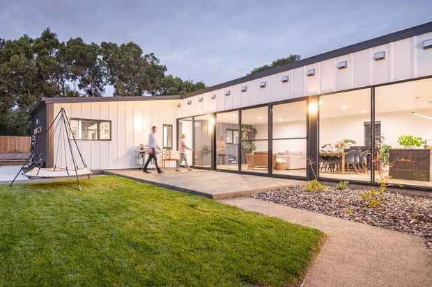 Single-storey contemporary building viewed from a landscaped outdoor area, with a long low roofline, pale vertical metal cladding, extensive black-framed floor-to-ceiling glazing, and a covered outdoor dining or living area. A paved path, gravel drainage bed, lawn, planted raised beds, and a hanging outdoor swing are visible. Warm interior lighting creates an inviting evening atmosphere.