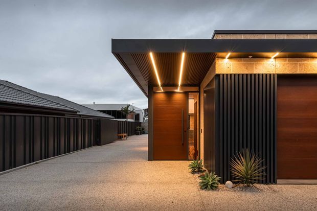 Contemporary residential entrance viewed from an exterior driveway, featuring a flat-roofed form with deep projecting eaves, dark vertical metal screening, warm timber-clad entry and garage doors, integrated linear lighting, and sculptural drought-tolerant planting in a gravel forecourt. A neighbouring pitched-roof dwelling is visible to the left.