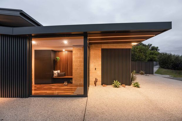 Contemporary single-storey house exterior at dusk, featuring a dark flat-roofed projecting canopy, black vertical ribbed cladding, extensive glazing, warm recessed lighting, and a textured pale concrete driveway or forecourt. Through the glazing, a warm timber-lined entry or living area is visible. Minimalist landscaping with sculptural low plantings complements the strong horizontal and vertical geometry.