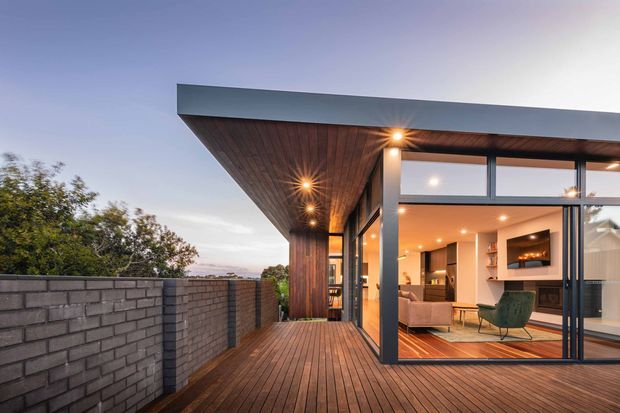 Contemporary single-level home viewed from a timber deck at dusk, featuring a broad flat roof with a deep timber-lined eave, floor-to-ceiling glazed living area, dark-framed windows and doors, and warm interior lighting. A grey masonry boundary wall and landscaped trees sit alongside the deck, while the visible interior includes a living room with a fireplace and built-in shelving.