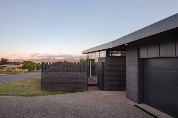 Exterior view of a contemporary single-storey home at dusk, featuring a low-slung flat roof, dark brick walls, charcoal vertical cladding, large glazed entry openings, and a wide dark garage door. A brick privacy wall and vertical metal screening define the entry court, with suburban surroundings and distant hills beyond.
