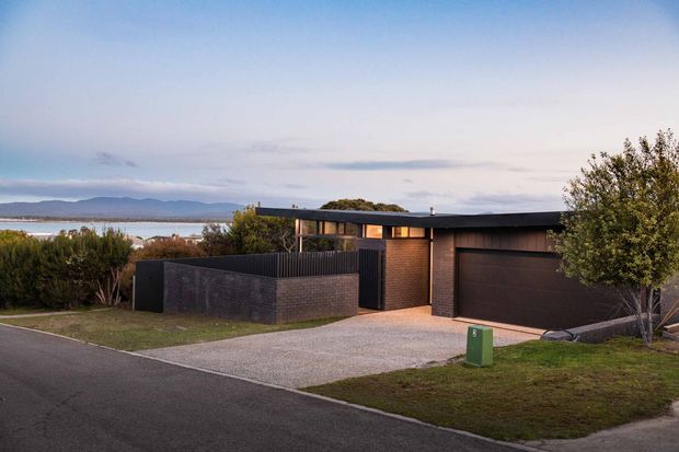 A low-profile contemporary single-storey house viewed from the street, with dark brick walls, a broad flat roof, integrated double garage, enclosed front courtyard, and a pale gravel driveway. The property overlooks a body of water and distant hills, with mature planting framing the facade.