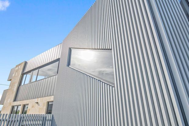 Upward-looking exterior view of a contemporary multi-level building with a strongly sculptural facade. Tall grey vertical ribbed metal cladding dominates the elevation, contrasted by large dark-framed windows and sections of pale stone cladding. The clear blue sky and dramatic perspective emphasize the building’s height and sharp geometric composition.
