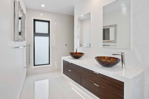 Bright contemporary double-vanity bathroom with a floating dark timber vanity, white solid-surface benchtop, vessel basins, wall-mounted chrome tapware, large frameless mirrors, glossy light flooring and walls, and a tall frosted window. A decorative framed mirror is also visible on the side wall.