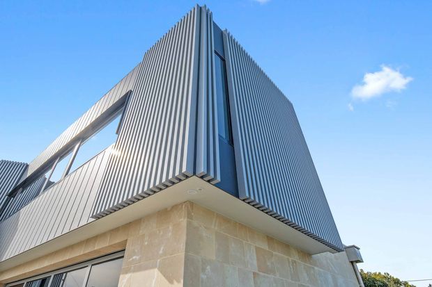 Upward-looking exterior architectural photograph of a sharply articulated contemporary building corner. The upper level is wrapped in closely spaced vertical ribbed metal cladding with narrow dark-framed glazing, while the lower facade uses large pale beige stone blocks. Strong geometric lines, a projecting upper volume, and a clear blue sky create a crisp, modern appearance.