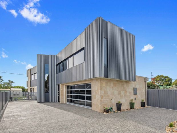 Contemporary two-storey residential building viewed from the driveway, with a strongly articulated corner form. The upper level is clad in vertically ribbed grey metal, while the lower level features pale natural stone masonry around a glazed sectional garage door. Additional dark-framed glazing and a tall narrow glazed slot bring light into the facade. A broad gravel driveway, metal fencing and gate surround the building.