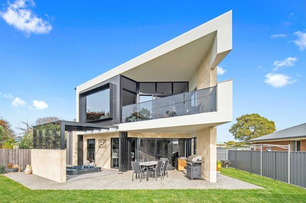 Bright exterior view of a contemporary two-storey residence with a dramatic asymmetrical pitched roof, white rendered surfaces, dark-framed glazing, a glass balcony balustrade, and a covered ground-floor outdoor dining and barbecue area. The home sits in a suburban landscaped yard with a paved terrace and boundary fencing.