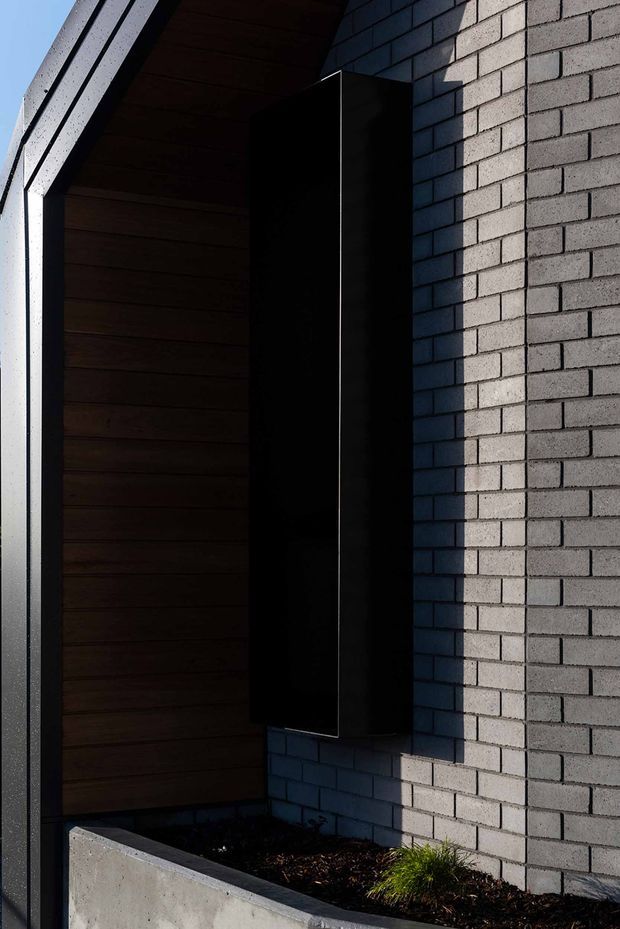 Close-up exterior architectural detail showing a dark recessed entry or porch framed by horizontal dark timber lining, a tall matte-black vertical door or panel, pale grey masonry brickwork, and a raw concrete planter at the base. Strong directional sunlight creates deep shadows and highlights the geometric, minimalist composition.