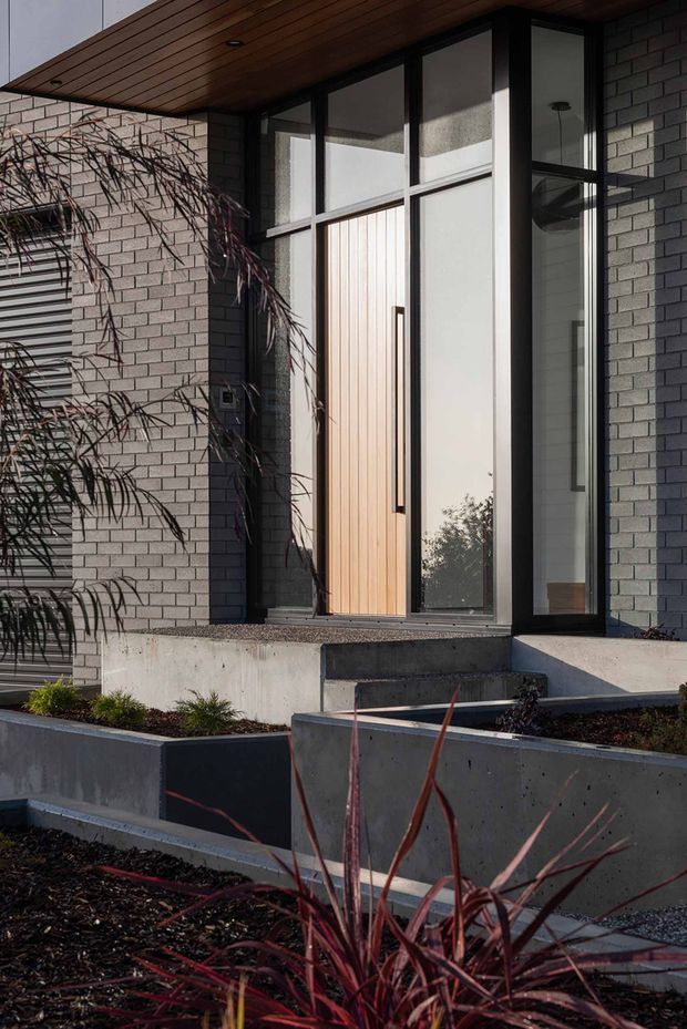 Close-up exterior view of a contemporary residential entry. The facade combines light grey brickwork with black-framed glazing and a tall vertical timber door beneath a projecting timber-lined soffit. Concrete steps, raised geometric garden beds, and drought-tolerant ornamental planting lead to the recessed front entrance.