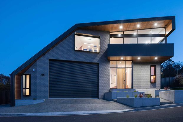 Contemporary two-storey residential home photographed at dusk, with a sharply angled roofline, dark brick facade, large charcoal garage door, timber-lined soffits, expansive upper-level glazing, and a projecting dark balcony. Concrete steps and raised planters form the front entry landscaping.